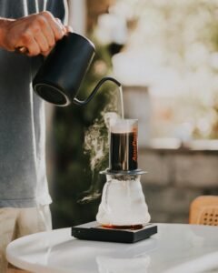 Hands pouring hot water into an Aeropress coffee maker on a sunny day, creating steam.